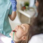 Crop faceless female dentist in uniform holding mouth mirror while preparing for inspecting female patient teeth with assistant holding tube suction in patient mouth